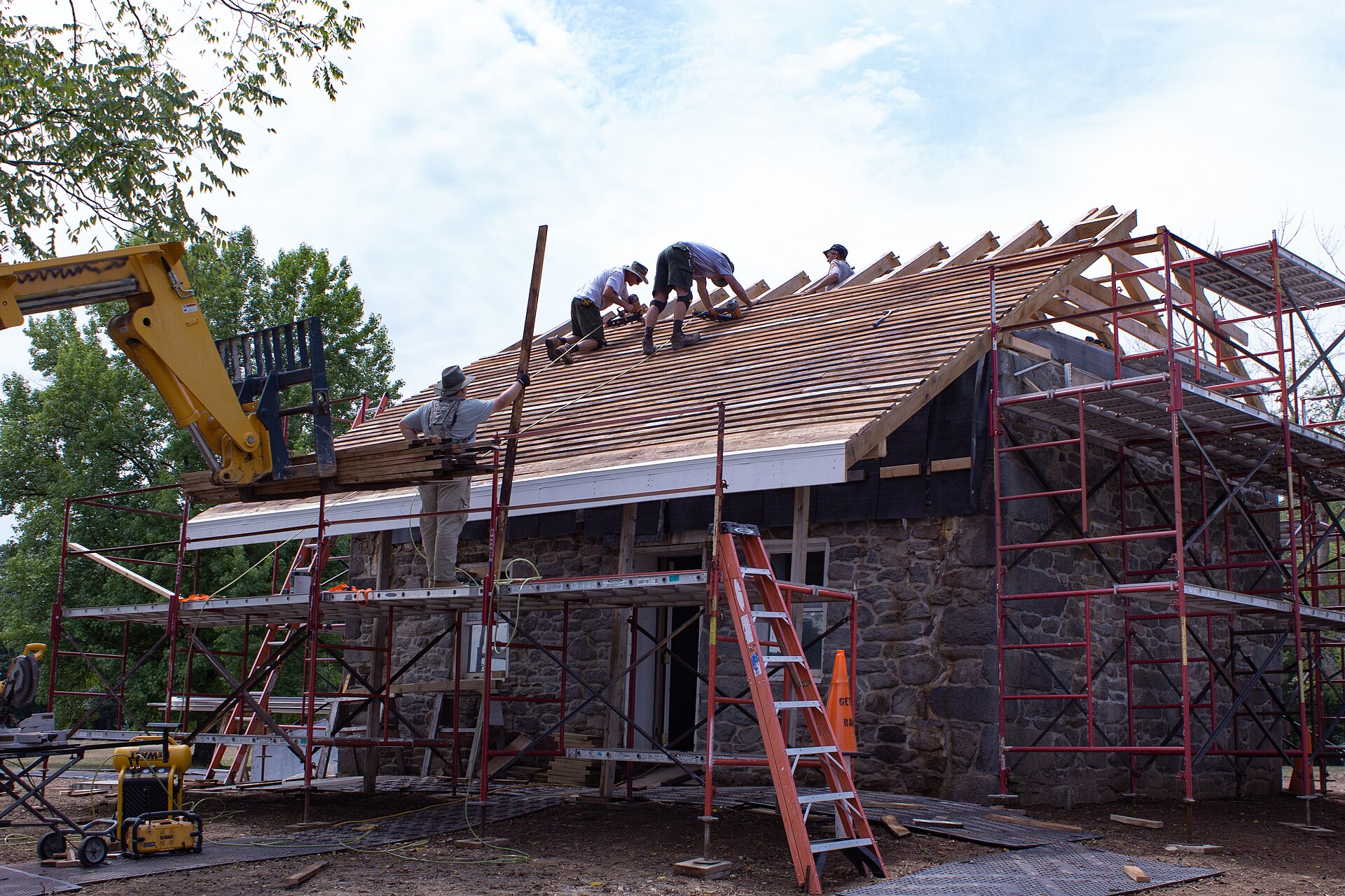 A roofing crew installing cedar shingles near the ridge of a house, with material lifted into place by a telehandler